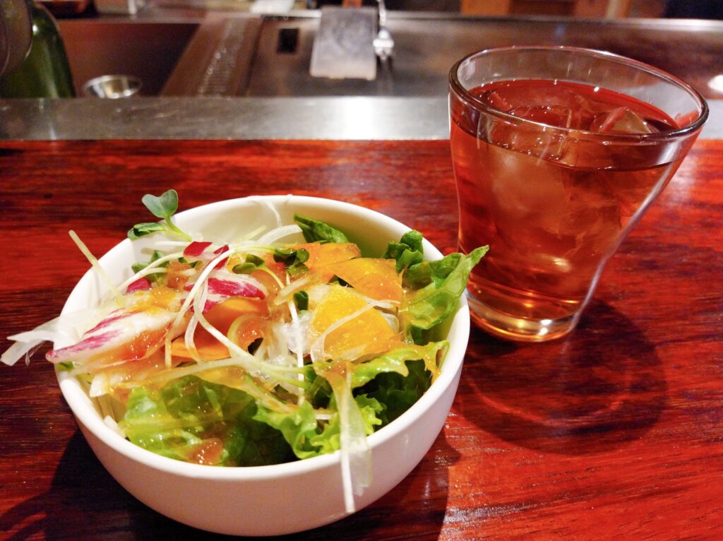 木目カウンターの上に小皿に入ったサラダとグラスに入ったウーロン茶が置かれている